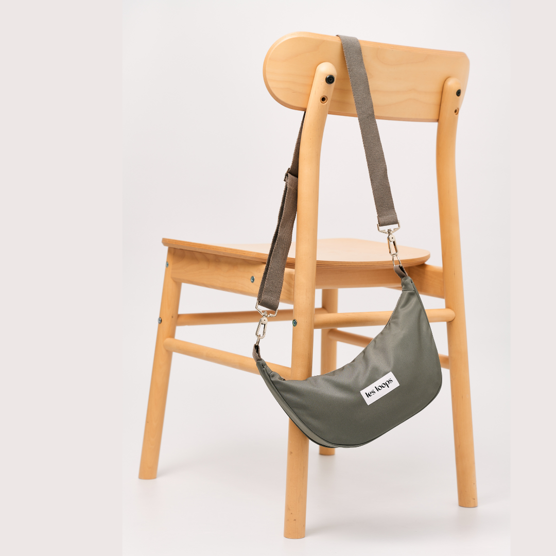 Die halbmondförmige Crossbody Bag Graugrün hängt lässig über der Schulter oder an einem Stuhl, der verstellbare Tragegurt ist abnehmbar